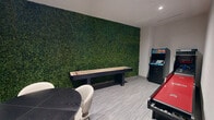 This well-appointed game room offers a dynamic and social atmosphere, thoughtfully designed for entertainment and leisure. The space is anchored by a striking floor-to-ceiling living green wall that adds a bold, natural focal point and a vibrant contrast to the room's neutral gray and white palette. Recessed lighting illuminates the space evenly, highlighting the light gray textured flooring that runs throughout.

The room is outfitted with an impressive array of recreational amenities. A full-length shuffleboard table is positioned along the green wall, while a red billiards table commands attention at the center of the space. A dedicated alcove houses a classic arcade cabinet and a skee-ball lane, offering retro gaming entertainment. A round black table surrounded by contemporary white upholstered chairs provides a comfortable seating area for socializing or casual gaming.

Framed vintage-style artwork and game-themed prints decorate the walls, contributing to the room's playful yet sophisticated character. A sleek black sideboard adds storage and refinement. Black-framed glass double doors connect the space to an adjacent area, enhancing the open and accessible flow. This game room is a comprehensive recreational retreat designed to accommodate a variety of activities and group sizes.