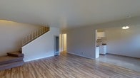 This two-story home offers a clean, functional layout designed for comfortable everyday living. The main level features an open living area finished with light-toned hardwood flooring that extends throughout, lending warmth and continuity to the space. Neutral gray walls and white trim create a bright, understated aesthetic that allows for versatile furnishing options. A staircase with a metal railing rises from the main living area, providing access to the upper floor and serving as a natural focal point within the room. A wall-mounted heating unit is positioned near the base of the stairs, highlighting the practical character of the home. Adjacent to the living area, a compact kitchen is clearly visible and features white cabinetry, a white range, and countertop workspace — offering an efficient and tidy cooking environment. A doorway near the base of the stairs suggests additional rooms or utility spaces on the main level. The upper floor adds further living space, making this home well-suited for those seeking a multi-level layout. Overall, the space presents a straightforward and move-in-ready interior with consistent finishes and a light, airy feel throughout.