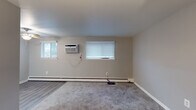 This single-floor unit offers a practical and comfortable layout across several well-defined living areas. The main living space is generously sized, featuring neutral-toned walls, plush wall-to-wall carpeting, and multiple windows fitted with white blinds that allow natural light to filter in. A wall-mounted air conditioning unit and baseboard heating provide year-round climate control, ensuring comfort in all seasons. The entry area opens into this open living space, which is accented by soft, neutral gray tones and clean white trim throughout.

The kitchen is a functional highlight of the unit, showcasing warm honey-toned wood cabinetry, granite-look countertops, a white tile backsplash, and a stainless steel sink. A freestanding electric range with a ventilation hood completes the cooking area, offering ample counter space for meal preparation.

The bedroom is a quiet, carpeted retreat with a window overlooking greenery, a baseboard heater, and double louvered sliding closet doors providing generous storage. The neutral color palette and soft carpeting lend a calm, restful ambiance to the room.

Overall, the space presents a clean, functional aesthetic with consistent neutral tones, practical finishes, and thoughtful climate management features throughout.