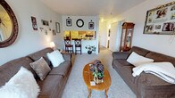 This single-floor unit features a well-organized layout that maximizes its comfortable footprint. The living room serves as the heart of the space, offering room for a full seating arrangement centered around a coffee table, with plush carpeting underfoot that adds warmth to the area. Neutral gray walls keep the space feeling open and versatile.

A defining architectural feature is the pass-through counter connecting the living area to the kitchen, which enhances the open feel while maintaining a distinct cooking zone. The kitchen is equipped with wood-toned cabinetry, a white refrigerator, and tiled backsplash, with bar-height seating along the counter providing a casual dining option.

A hallway visible from the living room leads to the bedroom and bathroom, offering a practical and private separation of sleeping quarters from the main living space. The consistent carpeting throughout the living areas contributes to a cohesive, cozy ambiance. Overall, the unit presents a functional and comfortable single-level layout well-suited for everyday living.