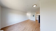 The Theadore Studio in Tacoma, WA offers a thoughtfully organized single-floor layout that makes efficient use of its compact footprint. The open living area is finished with light-toned hardwood flooring and crisp white walls, creating a clean, bright atmosphere. A large window allows natural light to fill the space while offering a view of surrounding greenery, and a wall-mounted HVAC unit provides climate control. Flush-mount ceiling lighting illuminates the interior throughout. The kitchen is positioned at one end of the unit and features white cabinetry, a countertop workspace, a sink, and a full-size refrigerator, providing all the essential amenities for everyday living. The bathroom is accessed via a sliding door and includes wood-look flooring that carries through from the main space, a walk-in shower with a curtain rod, a toilet, and a vanity with a light stone countertop and white cabinetry. The unit's clean, minimalist design and neutral color palette create a versatile and functional living environment well-suited for single occupancy in the Tacoma, WA area.
