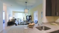 This single-floor space showcases an open-concept design that seamlessly connects the kitchen, dining, and living areas into one cohesive, flowing layout. The kitchen features granite countertops with a stainless steel sink, complemented by dark cabinetry that provides a striking contrast against the warm, light yellow walls. A modern square pendant light with a clean geometric frame hangs above, serving as a focal point that bridges the kitchen and dining zones.

The dining area transitions naturally into the living space, which is furnished with contemporary seating and a glass-topped coffee table. A dark media unit anchors the living area, adding structure and storage. Large windows dressed with sheer white curtains allow generous amounts of natural light to fill the space, enhancing its open and airy feel.

A glimpse through an interior doorway reveals an adjacent room, suggesting a thoughtfully connected floor plan. The overall ambiance is modern and polished, with a cohesive color palette of soft neutrals, dark furnishings, and metallic accents that lend the space a sophisticated yet comfortable character.