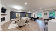 This well-appointed single-floor space offers a generously sized open-concept layout that seamlessly connects three distinct functional zones. The living lounge area is anchored by a stone-surround fireplace with a wood mantel and a wall-mounted television, complemented by comfortable neutral-toned seating arranged over a textured area rug. Recessed lighting and a stylish pendant fixture illuminate the space, while bold slate-blue accent walls add depth and visual contrast against the crisp white ceilings and trim throughout.

Flowing naturally from the lounge, the dining area features a large rectangular wood table with upholstered seating for multiple guests, positioned beneath a striking oversized wall clock. Adjacent accent chairs near a wide window provide a relaxed nook with views of lush greenery outdoors, filling the space with natural light.

The kitchen is outfitted with stainless steel appliances including a gas range, refrigerator, and dishwasher, paired with rich dark wood cabinetry, white subway tile backsplash, and open shelving. A substantial island with a white quartz countertop offers bar seating beneath elegant pendant lights. Durable wood-look flooring runs continuously throughout, unifying the entire space with a warm, contemporary aesthetic.
