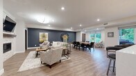 This well-appointed single-floor space offers a generously sized open-concept layout that seamlessly connects three distinct functional zones. The living lounge area is anchored by a stone-surround fireplace with a wood mantel and a wall-mounted television, complemented by comfortable neutral-toned seating arranged over a textured area rug. Recessed lighting and a stylish pendant fixture illuminate the space, while bold slate-blue accent walls add depth and visual contrast against the crisp white ceilings and trim throughout.

Flowing naturally from the lounge, the dining area features a large rectangular wood table with upholstered seating for multiple guests, positioned beneath a striking oversized wall clock. Adjacent accent chairs near a wide window provide a relaxed nook with views of lush greenery outdoors, filling the space with natural light.

The kitchen is outfitted with stainless steel appliances including a gas range, refrigerator, and dishwasher, paired with rich dark wood cabinetry, white subway tile backsplash, and open shelving. A substantial island with a white quartz countertop offers bar seating beneath elegant pendant lights. Durable wood-look flooring runs continuously throughout, unifying the entire space with a warm, contemporary aesthetic.