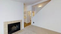 This two-story residence offers a thoughtfully arranged layout with distinct living zones connected by an open staircase. The main living area is finished with neutral wall-to-wall carpet and anchored by a wood-burning fireplace framed in light ceramic tile, providing a functional focal point for the room. Clean white walls and a crisp, minimal aesthetic give the space a bright, airy feel throughout.

Adjacent to the living area, the transition to the kitchen and surrounding spaces is marked by a shift to hardwood-style flooring, adding visual contrast and defining the separate functional zones. The kitchen area is partially visible through an open archway, featuring white cabinetry and overhead lighting that contributes to its bright, clean character.

The open staircase rising along the rear wall leads to the second floor, with a wall-mounted sconce providing warm accent lighting along the stairwell. The open sightlines between floors create a sense of vertical spaciousness. Overall, the space maintains a consistent, neutral palette throughout, offering a clean and versatile interior suited for a range of furnishing styles.