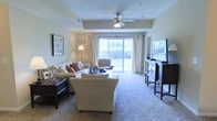 This single-floor residence offers a thoughtfully arranged living space designed with comfort and functionality in mind. The living room serves as the heart of the home, centered around a generous seating arrangement with a plush sofa, accent chair, and a cushioned armchair positioned to take full advantage of the natural light that pours in through large sliding glass doors. The doors open to an outdoor view, flooding the interior with brightness and creating a seamless connection between the indoors and outside. Soft, neutral-toned carpeting runs throughout the living area, lending a warm and inviting feel underfoot. A ceiling fan overhead provides both practical ventilation and a classic finishing touch to the space. The room is further appointed with tasteful furnishings, including dark-wood side tables and a media console that grounds the space with contrast against the light walls. Decorative wall art adds personality without overwhelming the calm, understated aesthetic. Recessed lighting and a floor lamp provide layered illumination for versatile use at any time of day. Overall, the space presents a clean, comfortable, and well-lit environment suited for everyday living and relaxed entertaining.