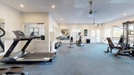 Located at 1345 S Kolb Rd in Tucson, Arizona, this fitness center offers a dedicated exercise space designed to support a well-rounded workout routine. The single-floor layout provides a comfortable and functional environment, with blue carpet flooring throughout that defines the workout zones clearly.

The cardio area is well-stocked with multiple treadmills, an elliptical trainer, and a stationary bike, all positioned to take advantage of the natural light streaming in through large windows along the perimeter. A multi-station cable strength machine anchors the free weights and strength training zone, providing versatile options for resistance-based workouts.

The interior design strikes an energetic tone, featuring bold athletic murals with teal, orange, and white geometric accents that add visual dynamism to the space. Ceiling fans — both a modern, sleek model and a pendant-style fan — keep air circulating throughout the room. Recessed lighting supplements the ample natural light, ensuring a bright atmosphere at any time of day.

A wall-mounted flat-screen TV and a decorative inset display shelf with geometric art contribute to the polished aesthetic. Large mirrors along one wall visually expand the space and assist with form during exercise. Glass-paneled doors provide direct outdoor access, enhancing ventilation and connectivity to the surrounding environment.