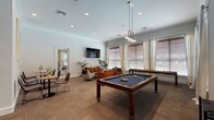 This single-floor amenity lounge is thoughtfully designed to offer a versatile and welcoming social environment. The space is anchored by a full-size billiards table at its center, illuminated by a sleek pendant light fixture that adds a modern, polished touch to the room. Large windows dressed with wooden blinds and flowing curtains flood the interior with natural light, creating a bright and inviting atmosphere throughout.

The lounge area features a curated seating arrangement of rich cognac leather armchairs and a plush accent chair, gathered around a low-profile coffee table. A wall-mounted flat-screen TV and a warm wooden credenza with decorative potted palms complete the relaxed, living-room-style setting. Framed koi fish artwork adorns one wall, adding an artistic focal point to the space.

A casual dining nook with bistro-style tables and chairs offers a more intimate setting for smaller gatherings. Neutral-toned carpet tiles cover the floors throughout, contributing to the space's understated elegance.

A connected kitchenette is fully equipped with stainless steel appliances, including a microwave, under-counter refrigerator, dishwasher, and sink, all set against a striking floor-to-ceiling textured tile backsplash with clean white countertops. French doors connect to an adjacent room, further enhancing the space's open and functional layout.