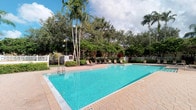This single-floor property is highlighted by a generously sized outdoor swimming pool that serves as the centerpiece of its exterior amenity space. The pool features clear, turquoise water and is framed by a well-maintained tile and paver deck, offering ample room for relaxation and leisure. Surrounding the pool area, a collection of lounge chairs provides comfortable seating for residents or guests seeking to enjoy the outdoors. The landscape is lush and tropical in character, with tall, swaying palm trees and a dense backdrop of mature trees and shrubs that create a sense of privacy and seclusion. A white perimeter fence defines the space while maintaining an open, airy feel. The overall ambiance is tranquil and resort-like, evoking the warmth and greenery characteristic of a South Florida setting. The natural tree canopy filters the sunlight and adds to the verdant, inviting atmosphere of the outdoor area, making the pool deck a standout feature of the property.