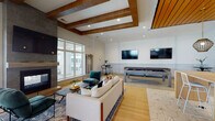 Located at 4027 Watercraft Ferry Avenue in Wilmington, NC, this contemporary single-floor community clubhouse showcases upscale design with a seamless blend of warm wood accents and clean modern finishes throughout. The main lounge area is anchored by a dramatic floor-to-ceiling stone fireplace with a mounted flat-screen television, complemented by exposed wood beam ceilings that add architectural depth and warmth. Comfortable seating arrangements, herringbone-patterned hardwood flooring, and large windows that invite ample natural light create an inviting social atmosphere. A billiards table and additional wall-mounted televisions offer varied entertainment options. Adjacent to the lounge, a dedicated wet bar and kitchen space features bold geometric hex-tile flooring, a wood-slat ceiling, pendant lighting, a built-in sink with dark countertops, and a long communal island with bar seating — ideal for gatherings and events. A private meeting or conference room offers glass partition walls with black steel framing, providing visual connectivity to the main space while maintaining a focused work environment. An interior staircase with sleek black metal railings connects areas and adds to the architectural character. Throughout, large windows provide views of the surrounding community, enhancing the bright, open ambiance of this well-appointed space.