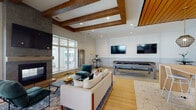 Located at 4027 Watercraft Ferry Avenue in Wilmington, NC, this contemporary single-floor community clubhouse showcases upscale design with a seamless blend of warm wood accents and clean modern finishes throughout. The main lounge area is anchored by a dramatic floor-to-ceiling stone fireplace with a mounted flat-screen television, complemented by exposed wood beam ceilings that add architectural depth and warmth. Comfortable seating arrangements, herringbone-patterned hardwood flooring, and large windows that invite ample natural light create an inviting social atmosphere. A billiards table and additional wall-mounted televisions offer varied entertainment options. Adjacent to the lounge, a dedicated wet bar and kitchen space features bold geometric hex-tile flooring, a wood-slat ceiling, pendant lighting, a built-in sink with dark countertops, and a long communal island with bar seating — ideal for gatherings and events. A private meeting or conference room offers glass partition walls with black steel framing, providing visual connectivity to the main space while maintaining a focused work environment. An interior staircase with sleek black metal railings connects areas and adds to the architectural character. Throughout, large windows provide views of the surrounding community, enhancing the bright, open ambiance of this well-appointed space.
