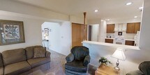 This single-floor space features an open-concept layout that connects the living area and kitchen, creating a cohesive and functional environment. The living area is carpeted and comfortably accommodates seating, including a cushioned sofa and an oversized armchair, contributing to a relaxed, cozy ambiance. Neutral-toned walls and recessed ceiling lighting provide a clean, well-lit atmosphere throughout the space.

A half-wall or breakfast bar separates the living area from the kitchen, maintaining visual openness while defining each zone. The kitchen is finished with warm wood cabinetry, white appliances including a range and refrigerator, and light-toned countertops. An under-cabinet mounted appliance and additional upper cabinetry offer practical storage solutions.

A wooden newel post near the kitchen area hints at an architectural transition point within the layout, adding a subtle traditional detail to the otherwise straightforward design. A sliding wood-panel closet door is visible near the living area, providing convenient storage. A side table with a lamp and small decorative accents complete the space, adding warmth to the overall setting.