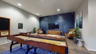 Located at 73 Brownstone Way in Englewood, NJ, this single-floor community lounge is designed to offer residents a well-rounded social and recreational experience. The space is thoughtfully laid out to accommodate both lively gatherings and relaxed downtime.

At the heart of the lounge, a full-size shuffleboard table commands attention, anchored atop a bold blue geometric-patterned area rug that adds a striking visual element to the hardwood-style flooring throughout. Facing the shuffleboard is a comfortable leather sofa accompanied by a marble-top coffee table, creating an inviting seating area ideal for casual conversation or relaxation.

A deep navy blue accent wall serves as a dramatic focal point, featuring a wall-mounted flat-screen television flanked by warm cognac leather wingback chairs and a wooden media console. Potted greenery is distributed throughout the space, adding natural freshness to the contemporary interior.

A dedicated game corner showcases a large wall-mounted Scrabble board alongside a compact bistro-style table with stools, while a collection of board games is neatly stacked nearby, encouraging interactive play. Large windows along one wall allow natural light to flow in, offering views of the surrounding brick exterior streetscape.

Black-framed glass doors provide a modern architectural detail and connect the lounge to adjacent areas, enhancing the open, accessible feel of the space.
