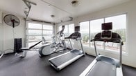 Located at 3261 SW Avalon Way in Seattle's West Seattle neighborhood, this well-equipped fitness center offers a comprehensive workout environment on a single floor. The space is defined by its striking floor-to-ceiling glass garage doors, which flood the interior with natural light and frame sweeping views of the surrounding Seattle skyline and greenery — creating an energizing atmosphere for exercise.

The fitness center is outfitted with a full suite of cardio equipment, including multiple treadmills and Precor elliptical machines positioned to take advantage of the panoramic views. The free weights area features a complete dumbbell rack alongside adjustable benches and large wall-mounted mirrors that enhance the sense of space. Medicine balls, foam rollers, and storage baskets provide additional functional training options.

The interior design incorporates bold yellow and white vertical stripes along the walls, adding a vibrant and modern aesthetic contrast to the dark rubber flooring, which is both practical and durable. Ceiling-mounted fans and a wall-mounted mini-split HVAC unit ensure year-round climate comfort. The space is completed by a keyed entry door with posted fitness center rules, offering a secure and organized environment for users.
