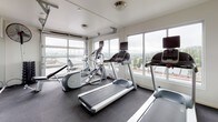 Located at 3261 SW Avalon Way in Seattle's West Seattle neighborhood, this well-equipped fitness center offers a comprehensive workout environment on a single floor. The space is defined by its striking floor-to-ceiling glass garage doors, which flood the interior with natural light and frame sweeping views of the surrounding Seattle skyline and greenery — creating an energizing atmosphere for exercise.

The fitness center is outfitted with a full suite of cardio equipment, including multiple treadmills and Precor elliptical machines positioned to take advantage of the panoramic views. The free weights area features a complete dumbbell rack alongside adjustable benches and large wall-mounted mirrors that enhance the sense of space. Medicine balls, foam rollers, and storage baskets provide additional functional training options.

The interior design incorporates bold yellow and white vertical stripes along the walls, adding a vibrant and modern aesthetic contrast to the dark rubber flooring, which is both practical and durable. Ceiling-mounted fans and a wall-mounted mini-split HVAC unit ensure year-round climate comfort. The space is completed by a keyed entry door with posted fitness center rules, offering a secure and organized environment for users.