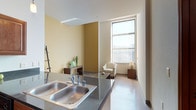This single-floor unit combines modern finishes with a practical, open layout. The kitchen serves as a central hub, featuring rich wood cabinetry, dark granite countertops, and a full suite of stainless steel appliances including a refrigerator, range, and built-in microwave. A peninsula with an integrated double sink and bar seating connects the kitchen to the living area, where polished concrete floors and tall, vertical-blind-dressed windows flood the space with natural light and views of the surrounding area. The living room offers a comfortable, open feel with warm yellow-toned walls that complement the modern furnishings.

The unit includes two carpeted bedrooms, each with ceiling fans and generous windows that bring in daylight. Both bedrooms are served by sliding closet doors providing ample storage, with one room featuring an additional walk-in closet with wire shelving. The two bathrooms are finished with dark vanity cabinetry and stone-look tile flooring. One bathroom includes a walk-in shower with a curtained surround, while the second is a half bath. Throughout the unit, clean lines and a neutral palette create a cohesive, contemporary ambiance.