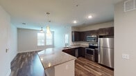 Located at 751 N Main St in Mansfield, Texas, this well-appointed single-story residence combines modern finishes with a functional, open layout. The heart of the home is the open-concept kitchen and living area, where rich dark cabinetry is paired with granite countertops, a large kitchen island, and a full suite of stainless steel appliances including a refrigerator, range, microwave, and dishwasher. Pendant lighting over the island adds a contemporary touch, while wood-look flooring extends through the main living spaces, creating a warm, cohesive aesthetic. Large windows allow natural light to fill the open living and dining areas, which flow seamlessly from the kitchen. The bedrooms are carpeted for comfort and include ceiling fans and window blinds. The primary suite features direct access to a private bathroom equipped with a glass-enclosed shower, dark wood vanity, and granite countertop, as well as a walk-in closet with built-in shelving. A secondary bathroom offers a soaking tub and shower combination with floor-to-ceiling tile surround, complemented by matching dark cabinetry and granite finishes. The unit's neutral color palette and consistent design language throughout create a clean, modern ambiance.