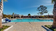 Located at 214 Kingsmeadow Cir in Belville, NC, this single-level outdoor amenity complex offers a thoughtfully designed resort-style retreat. At its centerpiece sits a generously sized swimming pool with crystal-clear blue water, complemented by a separate spa with bubbling water features. The pool deck is finished in elegant stamped concrete pavers, providing ample space for sunbathing and socializing. Multiple rows of cushioned lounge chairs are arranged throughout the deck, offering residents a variety of spots to relax in the North Carolina sun. A series of modern white pergolas with slatted roofing provides shaded seating areas along one side of the pool, while warm-toned horizontal wood privacy screens frame the perimeter, creating a sense of enclosure and sophistication. A dedicated social courtyard features a central fire pit surrounded by Adirondack-style chairs, ideal for evening gatherings. Tall palm trees, ornamental grasses, and carefully curated plantings lend a coastal, tropical ambiance to the entire space. A well-appointed clubhouse building with large windows, a metal roof, and a covered outdoor porch anchors one end of the complex. Additional amenity structures, including what appears to be restroom and storage facilities, round out this comprehensive and well-maintained outdoor living destination.