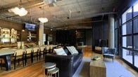 This single-floor space showcases a compelling industrial-chic design that blends raw architectural elements with warm, inviting furnishings. The exposed concrete ceiling highlights an open network of ducts and conduit, lending an authentic urban loft character throughout. Structural concrete columns punctuate the open floor plan, reinforcing the space's industrial roots while adding a sense of vertical scale.

Warm hardwood flooring runs throughout, providing a rich contrast to the cooler tones of the ceiling above. A central lounge area anchors the space with a substantial leather sofa, eclectic accent chairs, and a rustic solid-wood coffee bench, creating a comfortable and stylish focal point. A cluster of globe pendant lights adds a soft, sculptural lighting feature overhead.

Adjacent to the lounge, a long communal dining or gathering table lined with wooden chairs accommodates larger groups, making the space well-suited for events or collaborative use. Dark vertical slat paneling and dramatic black curtains define a more intimate zone toward the rear, adding depth and visual contrast. A bar or service area is visible in the background, further enhancing the space's versatility. The overall ambiance is sophisticated yet relaxed, blending modern design sensibilities with a distinctly urban, loft-inspired character.