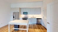 This thoughtfully designed one-bedroom, one-bathroom residence at Ascot in Royal Palm Beach, Florida offers a clean, modern aesthetic throughout its single-floor layout. The open-concept living and kitchen area is anchored by light wood-look flooring that flows seamlessly throughout the main living spaces. The kitchen is a standout feature, showcasing contemporary light gray cabinetry that extends to the ceiling, a white subway tile backsplash, sleek light countertops, and a full suite of stainless steel appliances including a French door refrigerator, range, microwave, and dishwasher. A kitchen island provides additional prep and dining space. The living area is bright and airy, with a ceiling fan overhead and sliding glass doors fitted with vertical blinds that open to an outdoor view, allowing ample natural light to fill the room. A convenient in-unit stacked washer and dryer is tucked neatly behind a louvered closet door near the kitchen. The bedroom is generously sized, featuring plush carpeting, a ceiling fan, and a walk-in closet with dual shelving rods. The bathroom is finished with large-format tile flooring, an extended vanity with light countertops, and a tiled walk-in shower.