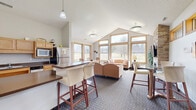 Located in Champlin, Minnesota, this single-floor space seamlessly combines a kitchen and living room into a cohesive, open-plan layout designed for comfort and functionality. The living area is anchored by a striking floor-to-ceiling stacked stone fireplace with a mounted flat-screen TV, serving as a natural focal point. Soaring vaulted ceilings amplify the sense of openness, while an expansive bank of large windows with warm wood-toned frames floods the space with natural light and frames serene, pastoral views of the surrounding greenery. Patterned carpet in a cool, neutral tone adds texture and warmth underfoot throughout the living zone. A bar-height counter with barstool seating creates a natural transition between the living and kitchen areas, ideal for casual dining or entertaining. The kitchen is practical and well-appointed, featuring light maple cabinetry, dark countertops, tile flooring, and a full suite of stainless steel appliances including a refrigerator, dishwasher, microwave, and electric range. Pendant and flush-mount lighting provide layered illumination throughout. A patio area visible from the exterior rounds out this inviting, light-filled space.