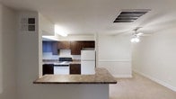 This single-floor residence features an open-concept layout connecting the kitchen and living area. The kitchen is well-appointed with rich, dark wood cabinetry providing ample upper and lower storage. Granite-style countertops extend along the workspace and continue onto a peninsula breakfast bar, offering a functional partition between the kitchen and the adjacent living space. White appliances, including a gas range and full-size refrigerator, complement the neutral color palette throughout. A ceiling-mounted light fixture illuminates the kitchen, while a large overhead HVAC vent ensures climate control. The adjoining living area is finished with light-toned carpeting and features a ceiling fan with integrated lighting, enhancing air circulation and ambient light. Crisp white walls and clean baseboard trim run throughout both spaces, contributing to a cohesive, well-maintained aesthetic. The open sightlines between the kitchen and living area create a sense of spaciousness, making the layout practical for everyday living and casual entertaining.