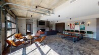 This single-floor amenity space blends industrial design with modern sophistication, creating a dynamic and versatile communal environment. Exposed concrete ceilings with visible ductwork and structural columns establish an urban, loft-like aesthetic throughout, while stone tile flooring and warm wood accents add texture and depth.

The main lounge area is anchored by a stone-surround fireplace flanked by built-in shelving and a wall-mounted TV, surrounded by plush seating in rich leather tones. A large communal dining table with bench seating occupies the central zone, complemented by a row of sleek gold-framed counter-height chairs along a bar-style ledge. Bold, colorful wall art adds vibrant personality throughout the space.

The fully equipped kitchen features dual refrigerators, generous counter space with a central island, warm wood cabinetry, stone tile flooring, and pendant lighting — designed for group gatherings and events.

A dedicated game room includes a regulation billiards table, a large-screen TV, framed artwork, and cozy lounge seating. Adjacent nook areas offer intimate seating with curated gallery walls and natural light from large windows.

A standout feature is the pair of full-height glass garage doors that open directly onto an outdoor pool deck, seamlessly connecting the interior with the exterior and flooding the space with natural light and open-air views.