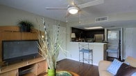 This single-floor space showcases an open-concept layout that connects the living area and kitchen in a cohesive, functional design. The living area features rich hardwood flooring, a ceiling fan with a light fixture overhead, and a textured ceiling that adds subtle dimension. A media console with warm wood tones anchors the space, while decorative elements like a tall ornamental grass arrangement in a green vase contribute a natural, organic touch.

The kitchen is defined by dark cabinetry that contrasts with a light-colored breakfast bar, creating a visually striking boundary between the two areas. Stainless steel appliances, including a refrigerator, are visible from the living space, emphasizing a modern aesthetic. A pass-through opening above the bar connects the kitchen to the living area, enhancing the open feel. A metal bar stool with a cushioned seat provides casual seating at the counter. Recessed lighting in the kitchen ceiling ensures the space is well-lit. Overall, the layout is practical and cohesive, with a neutral color palette and modern finishes creating a clean, contemporary ambiance throughout.