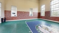 Located at 649 Alden St in Fall River, Massachusetts, this single-floor indoor sports and recreation facility is purpose-built for athletic training and leisure. The centerpiece of the space is a full-size indoor basketball court finished with a striking Boston Celtics-themed surface, complete with the iconic Celtics logo at center court and regulation markings rendered in green and blue interlocking sport flooring. A professional-grade adjustable basketball hoop with a clear backboard anchors one end of the court, while soaring ceilings with exposed wooden beams overhead provide the vertical clearance needed for full play. The court is surrounded by exposed brick wainscoting along the lower walls, which transitions to a warm beige upper wall finish, and a series of large, grid-paned windows flood the space with abundant natural light and views of the surrounding greenery. Adjacent to the main court, a dedicated indoor putting green offers a turf-lined practice lane for golf, complemented by overhead pendant lighting and padded training equipment nearby. Glass partition doors connect the court to additional areas of the facility. The overall ambiance is that of a high-end, well-appointed athletic complex designed to support a wide range of sports activities in a refined and comfortable indoor environment.