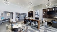 This single-floor clubhouse is designed with a sophisticated and contemporary aesthetic that creates a welcoming environment for socializing and relaxation. The main gathering area commands attention with soaring ceilings adorned by a striking geometric chandelier, lending the space an upscale, curated feel. The flooring features a bold geometric-patterned carpet that anchors the room and adds visual interest throughout.

A central highlight is the large wooden communal table surrounded by sleek leather counter-height chairs, ideal for casual meetings or informal gatherings. Flanking this area is a built-in shelving unit with warm wood tones that frames a fireplace and a mounted television, offering a cozy focal point for the lounge. Thoughtfully placed artwork and decorative accessories contribute to the refined ambiance.

Intimate seating clusters with plush upholstered chairs and round accent tables are arranged throughout the space, providing comfortable nooks for smaller groups. Recessed lighting and an open layout ensure the area remains bright and airy. An adjacent corridor, visible through an arched doorway, suggests additional amenity spaces beyond, expanding the overall functionality of the clubhouse. The overall design strikes a balance between elegance and approachability, making it a standout communal destination.