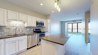 This well-appointed single-floor residence offers a thoughtfully designed open-concept layout that seamlessly connects the kitchen and living areas. The kitchen is a standout feature, showcasing white shaker-style cabinetry, a mosaic tile backsplash, dark countertops, and a full suite of stainless steel appliances including a refrigerator, range, microwave, and dishwasher. A peninsula with additional counter space provides both prep area and a casual dining option, while pendant lighting adds a warm, modern touch overhead. Light wood-style flooring runs throughout the main living areas, enhancing the bright and airy feel. Large picture windows in the living and dining spaces frame views of the surrounding urban streetscape, filling the rooms with natural light. The bedrooms offer plush wall-to-wall carpeting, ceiling fans, and oversized windows that continue the theme of abundant natural light and city views. The bathroom is finished with large-format tile flooring, white cabinetry with a granite-style countertop, an undermount sink, and a well-lit vanity mirror. Overall, the space presents a clean, contemporary aesthetic with consistent neutral tones and quality finishes throughout.