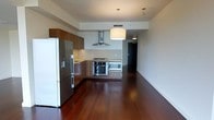 This single-floor space offers a sleek, modern design centered around an open-concept layout that seamlessly integrates the kitchen and living areas. The rich, dark hardwood floors extend throughout, lending warmth and elegance to the clean, contemporary aesthetic.

The kitchen is well-appointed with a full suite of stainless steel appliances, including a built-in wall oven, microwave, gas cooktop, and a large refrigerator. A stainless steel range hood serves as a functional focal point above the cooktop. The cabinetry features a warm wood-grain finish with a modern flat-front design, complemented by light countertops that provide a balanced contrast. Pendant lighting hangs from the ceiling, offering both task illumination and a stylish design element that enhances the open atmosphere.

The living area benefits from the open floor plan, creating a generous and flexible space that flows naturally from the kitchen. The neutral wall tones and clean ceiling lines contribute to an airy, uncluttered ambiance. A hallway visible in the background suggests additional rooms or storage areas beyond the main living space. Overall, the space presents a polished, move-in-ready environment with thoughtful finishes and a cohesive modern sensibility throughout.