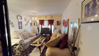 This single-floor space centers around a thoughtfully decorated living room that balances comfort and style. The room features two generously sized, cushioned sofas in a warm beige tone, accented with deep crimson throw pillows that add a rich contrast. A classic coffee table with ornate legs anchors the seating arrangement, resting atop a detailed, patterned area rug that adds warmth and texture to the floor.

A flat-screen television is positioned near a window dressed in dramatic burgundy drapes with gold tiebacks, which serve as a bold focal point against the neutral walls. The window allows natural light to filter into the space while the layered curtains provide privacy and a touch of elegance.

Decorative elements are abundant throughout, including wall-mounted artwork, framed photographs, ornamental plates, and tall decorative branches that add vertical interest. A multi-arm decorative floor lamp contributes both ambient lighting and visual character. The textured ceiling adds subtle architectural detail. Overall, the space presents a richly layered, eclectic aesthetic with warm tones and a lived-in, welcoming ambiance.