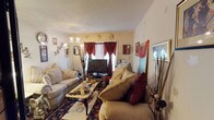 This single-floor space centers around a thoughtfully decorated living room that balances comfort and style. The room features two generously sized, cushioned sofas in a warm beige tone, accented with deep crimson throw pillows that add a rich contrast. A classic coffee table with ornate legs anchors the seating arrangement, resting atop a detailed, patterned area rug that adds warmth and texture to the floor.

A flat-screen television is positioned near a window dressed in dramatic burgundy drapes with gold tiebacks, which serve as a bold focal point against the neutral walls. The window allows natural light to filter into the space while the layered curtains provide privacy and a touch of elegance.

Decorative elements are abundant throughout, including wall-mounted artwork, framed photographs, ornamental plates, and tall decorative branches that add vertical interest. A multi-arm decorative floor lamp contributes both ambient lighting and visual character. The textured ceiling adds subtle architectural detail. Overall, the space presents a richly layered, eclectic aesthetic with warm tones and a lived-in, welcoming ambiance.