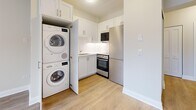 This efficiently designed one-bedroom, one-bathroom unit makes thoughtful use of its single-floor layout, offering a clean, modern aesthetic throughout. Light hardwood-style flooring runs continuously across all rooms, complemented by crisp white trim and neutral warm-toned walls that create a bright, cohesive interior.

The kitchen is compact yet highly functional, featuring white shaker-style cabinetry, a stainless steel refrigerator, built-in microwave, and a range. A standout feature is the dedicated in-unit laundry closet housing a stacked Bosch washer and dryer, adding exceptional convenience.

The living room is an open, sun-filled space with a sliding glass door that leads to a private balcony, offering elevated views of the surrounding urban landscape. Natural light pours in generously through the large glass door, creating an airy atmosphere.

The bedroom provides a comfortable retreat, complete with a sliding-door closet for storage and a calm, neutral palette. A connecting hallway leads efficiently to the full bathroom, which is finished in crisp white with a single vanity, mirrored medicine cabinet, toilet, and a soaking tub-shower combination. The hallway also provides access to additional closet space.

Overall, this unit delivers a clean, move-in-ready space with modern appliances, practical storage solutions, and desirable outdoor access.