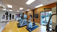 This fitness center offers a functional and inviting workout environment designed to support an active lifestyle. The space is anchored by warm hardwood flooring and striking exposed brick walls that lend a character-rich, industrial aesthetic to the room. A drop ceiling with large recessed panel lighting ensures the area is consistently well-lit, while generously sized windows along the exterior wall allow abundant natural light to flow in and provide views of the outdoors.

The cardio equipment area is outfitted with a row of Matrix-brand machines, including treadmills and an elliptical trainer, each positioned on protective black floor mats to preserve the hardwood beneath. A cable machine with a pulley system is mounted to the brick wall, adding a strength-training option to the space. A prominent informational board is displayed on the brick wall, providing members with guidelines and rules for facility use.

The layout is open and uncluttered, allowing for ease of movement between equipment. A secondary room or entry area is visible at one end, suggesting additional space or an accessible entrance. The overall ambiance is clean, practical, and welcoming — combining classic architectural details with modern fitness equipment for a motivating workout environment.