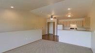 This single-floor space offers an open and functional layout designed for comfortable everyday living. The main living area features plush carpeting and warm neutral wall tones, creating a welcoming atmosphere. A ceiling fan with integrated lighting is centrally positioned, providing both air circulation and ambient light to the space. The living area transitions seamlessly into the kitchen, which is separated by a raised breakfast bar counter with a light-toned surface — ideal for casual dining or meal prep. The kitchen is equipped with light wood cabinetry, a white refrigerator, a range, and an over-the-range microwave or hood, all set on hard surface flooring that contrasts with the carpeted living zone. Recessed and flush-mount ceiling lighting throughout ensures the space remains well-lit. A doorway visible from the main living area leads to what appears to be a closet or additional room, suggesting practical storage solutions within the layout. The overall design is clean and straightforward, with a consistent warm beige palette throughout that lends the space a cohesive, understated character well-suited to a variety of furnishing styles.