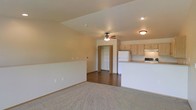 This single-floor space offers an open and functional layout designed for comfortable everyday living. The main living area features plush carpeting and warm neutral wall tones, creating a welcoming atmosphere. A ceiling fan with integrated lighting is centrally positioned, providing both air circulation and ambient light to the space. The living area transitions seamlessly into the kitchen, which is separated by a raised breakfast bar counter with a light-toned surface — ideal for casual dining or meal prep. The kitchen is equipped with light wood cabinetry, a white refrigerator, a range, and an over-the-range microwave or hood, all set on hard surface flooring that contrasts with the carpeted living zone. Recessed and flush-mount ceiling lighting throughout ensures the space remains well-lit. A doorway visible from the main living area leads to what appears to be a closet or additional room, suggesting practical storage solutions within the layout. The overall design is clean and straightforward, with a consistent warm beige palette throughout that lends the space a cohesive, understated character well-suited to a variety of furnishing styles.