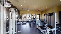 Located at 100 Village Way in Goleta, California, this single-level fitness center offers a comprehensive workout environment designed to support a wide range of exercise routines. The space is divided into distinct functional zones, each thoughtfully outfitted with professional-grade equipment.

The cardio area features multiple treadmills, elliptical trainers, and a stair climber, all positioned near large windows that allow natural light to fill the room and provide views of the surrounding landscaping. A bold athletic mural on one dark accent wall adds visual energy to this section of the facility.

The strength training zone is equipped with Precor cable and weight machines, a leg press, and a well-stocked dumbbell rack. Full-length mirrors line one wall, enhancing the sense of space and allowing users to monitor their form. A dedicated open floor area provides room for stretching and functional training, complemented by a wall-mounted television and yoga mats stored in a wicker basket nearby.

Throughout the facility, dark rubber flooring provides durability and safety, while recessed lighting and ceiling fans maintain a comfortable atmosphere. Windows on multiple walls invite natural light and connect the interior to the outdoors, contributing to an energizing and functional fitness environment.