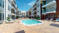 Located at 927 W Morgan St in Raleigh, NC, this outdoor courtyard amenity space offers a resort-inspired retreat at the heart of a multi-story residential community. The centerpiece is a generously sized swimming pool with crystal-clear blue water, framed by an expansive stone-paved patio finished in warm earth tones. The surrounding deck provides ample room for lounging, with an array of white chaise lounge chairs and shaded umbrella stations positioned throughout for comfort and relaxation.

The courtyard is enclosed by the building's multi-story facade, which features a lively mix of brick, white panels, and colorful accent details, along with private balconies that overlook the pool area. Decorative wrought iron fencing with ornamental detailing lines the perimeter, adding a classic and elegant touch to the outdoor space. Lush potted greenery and tropical plants are thoughtfully placed throughout, enhancing the vibrant, welcoming atmosphere.

A dedicated grilling area equipped with a charcoal grill offers an ideal setting for outdoor gatherings, while the brick accent walls and manicured plantings add texture and visual interest to the space. The entire area is bathed in natural sunlight, creating an open and energetic ambiance perfectly suited for outdoor recreation and social enjoyment in the heart of Raleigh.