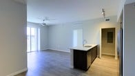 This single-floor space offers a clean, contemporary design centered around an open-concept living and kitchen area. The kitchen features a dark-cabinetry island with a stainless steel sink and light-toned countertop, providing both a functional workspace and a natural dividing point between the cooking and living zones. The living area is spacious and airy, finished with warm hardwood-style flooring that runs throughout, contributing to a cohesive and modern aesthetic. Neutral gray walls paired with bright white ceilings and trim create a crisp, versatile backdrop. A ceiling fan is centrally positioned in the living area, adding both practical airflow and a subtle design accent. Large sliding glass doors along one wall flood the space with abundant natural light and offer views to an outdoor area, enhancing the open and welcoming atmosphere. Recessed track lighting near the kitchen adds targeted illumination, complementing the overall well-lit environment. A doorway visible in the background hints at additional rooms beyond the main living space, while a secondary hallway opening suggests further utility areas. The overall ambiance is bright, minimal, and move-in ready, making it an adaptable space suited to a variety of uses.
