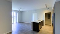 This single-floor space offers a clean, contemporary design centered around an open-concept living and kitchen area. The kitchen features a dark-cabinetry island with a stainless steel sink and light-toned countertop, providing both a functional workspace and a natural dividing point between the cooking and living zones. The living area is spacious and airy, finished with warm hardwood-style flooring that runs throughout, contributing to a cohesive and modern aesthetic. Neutral gray walls paired with bright white ceilings and trim create a crisp, versatile backdrop. A ceiling fan is centrally positioned in the living area, adding both practical airflow and a subtle design accent. Large sliding glass doors along one wall flood the space with abundant natural light and offer views to an outdoor area, enhancing the open and welcoming atmosphere. Recessed track lighting near the kitchen adds targeted illumination, complementing the overall well-lit environment. A doorway visible in the background hints at additional rooms beyond the main living space, while a secondary hallway opening suggests further utility areas. The overall ambiance is bright, minimal, and move-in ready, making it an adaptable space suited to a variety of uses.