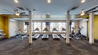 Located at 101 E Fourth St in Newport, Kentucky, this well-appointed fitness center offers a comprehensive range of exercise equipment and training zones across a single open floor. Large windows line the perimeter, flooding the space with natural light and providing views of the surrounding streetscape, while recessed lighting and ceiling fans maintain a comfortable atmosphere throughout. The cardio zone is anchored by a row of treadmills and elliptical machines positioned directly in front of the windows, complemented by upright stationary bikes for additional low-impact training options. A dedicated functional training area features a TRX suspension training rig mounted to the wall, along with a multi-station cable and weight machine for strength work. A spin cycling zone includes multiple bikes alongside a flat-screen TV, creating an engaging environment for indoor cycling sessions. Throughout the space, racks are stocked with stability balls, medicine balls, resistance bands, foam rollers, and yoga mats, supporting a wide variety of workout styles. Durable patterned carpet tiles cover the floors, while accent walls in bold yellow add visual energy to the space. Glass partition walls connect the fitness center to an adjacent lounge area, contributing to an open, integrated feel. Exposed dark ceiling beams in select areas add an architectural character to the overall design.
