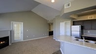 This single-floor space is defined by its striking vaulted ceilings, which lend an expansive, airy quality to the open-concept living and kitchen area. The living space is finished with neutral-toned carpet, while the kitchen transitions to hard flooring, creating a natural distinction between the two zones. The kitchen features a curved granite-style countertop peninsula, white cabinetry, and a full-size refrigerator, making it both functional and visually appealing. Built-in shelving is positioned adjacent to the kitchen, offering convenient storage and display space. The walls are finished in a soft gray tone throughout, contributing to a cohesive and contemporary aesthetic. A hallway visible from the main living area leads toward additional rooms, with dark cabinetry visible at its end suggesting a dedicated vanity or utility area. The ceiling-mounted light fixture and HVAC vents are integrated neatly into the vaulted ceiling. The overall design balances openness with practicality, creating a bright, well-organized living environment suited to everyday comfort.