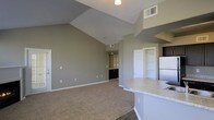 This single-floor space is defined by its striking vaulted ceilings, which lend an expansive, airy quality to the open-concept living and kitchen area. The living space is finished with neutral-toned carpet, while the kitchen transitions to hard flooring, creating a natural distinction between the two zones. The kitchen features a curved granite-style countertop peninsula, white cabinetry, and a full-size refrigerator, making it both functional and visually appealing. Built-in shelving is positioned adjacent to the kitchen, offering convenient storage and display space. The walls are finished in a soft gray tone throughout, contributing to a cohesive and contemporary aesthetic. A hallway visible from the main living area leads toward additional rooms, with dark cabinetry visible at its end suggesting a dedicated vanity or utility area. The ceiling-mounted light fixture and HVAC vents are integrated neatly into the vaulted ceiling. The overall design balances openness with practicality, creating a bright, well-organized living environment suited to everyday comfort.