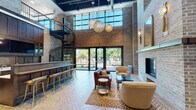 This two-story space masterfully combines industrial character with refined, hospitality-inspired design. The centerpiece of the main level is a dramatic double-height great room defined by exposed brick walls that soar upward to a wall of steel-framed windows, flooding the interior with abundant natural light. A striking black metal spiral staircase connects the two floors, anchoring one side of the room alongside a dark-wood bar with gold-toned barstool seating. Oversized globe pendant chandeliers hang at varying heights, adding warmth and visual drama overhead.

The lounge area is anchored by a floor-to-ceiling brick fireplace with a wood mantel, surrounded by plush seating arrangements on herringbone-patterned hardwood flooring. Large glass doors open directly to an outdoor patio, seamlessly extending the living space. A service counter with marble-veined countertops and rich wood cabinetry provides a bakery-style refreshment station.

A separate lounge room offers a more intimate setting, featuring patterned carpeting, sculptural gold velvet chairs, curated artwork, and elegant brass accent lighting. An entry vestibule with bold black-and-white marble checkerboard flooring, a coffered wood ceiling, globe sconces, and built-in bookshelves adds a distinguished, club-like arrival experience.