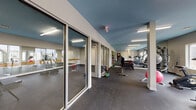 Located at 12191 Felch St in Holland, Michigan, this single-floor fitness facility offers a thoughtfully designed environment for a wide range of exercise activities. The main gym area is spacious and open, featuring durable rubber flooring throughout and a distinctive teal-painted ceiling that adds a modern, energetic aesthetic. The space is illuminated by overhead fluorescent lighting, complemented by natural light from windows along the perimeter.

The main floor is well-equipped with a comprehensive selection of free weights, including a full dumbbell rack, kettlebells of varying sizes, medicine balls, and cardio machines such as treadmills and stationary bikes. Large floor-to-ceiling mirrors line the walls, enhancing the sense of space and supporting form-focused training.

Glass-paneled sliding doors divide the facility into distinct zones, including two dedicated studio rooms. One studio features a wall-length mirror with a wooden ballet barre, making it ideal for dance, barre, or stretching classes. The second studio is a flexible open-floor space equipped with yoga mats on a storage rack, suited for group fitness or individual practice sessions.

Structural columns punctuate the main floor while maintaining an open and functional layout. The clean, neutral wall tones paired with the bold ceiling color create a motivating yet professional atmosphere throughout the facility.