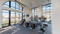Located at 16101 Murphy Ave in Irvine, CA, this state-of-the-art fitness center is designed to inspire and accommodate a wide range of workout routines. The space is defined by its dramatically high, double-height ceilings and an abundance of natural light streaming through floor-to-ceiling arched and grid-pane windows, offering views of the surrounding palm tree-lined landscape.

The cardio zone is generously equipped with treadmills, elliptical trainers, stair climbers, and stationary bikes, all arranged to maximize the open, airy feel of the space. Wall-mounted flat-screen televisions are positioned throughout for entertainment during workouts. Ceiling fans provide additional comfort.

The strength-training area features a multi-functional cable machine, a Smith machine, a full dumbbell rack with an adjustable weight bench, and additional resistance equipment, catering to both beginner and advanced fitness enthusiasts. Vibrant, colorful artwork accents the walls, adding an energetic touch to the environment.

Practical amenities include a dual stainless steel drinking fountain and water bottle refill station, conveniently located within the facility. The durable, patterned carpet flooring ties the space together, balancing functionality with a clean, contemporary aesthetic throughout this well-appointed fitness center.
