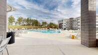 Located at 2525 Sycamore Farms Dr NW in Concord, NC, this impressive outdoor amenity complex delivers a resort-caliber experience designed for active recreation and relaxed leisure. At the heart of the space is a generously sized, freeform swimming pool with brilliantly clear blue water, lined on all sides by a broad concrete deck furnished with rows of white lounge chairs — ideal for sunbathing and socializing. Adjacent to the main pool is a separate shallow wading pool featuring in-water lounge chairs, offering a tranquil retreat for leisurely relaxation. A dedicated clubhouse structure anchors the complex, showcasing a welcoming architectural design with large windows, a covered porch, and tasteful exterior detailing that blends traditional and modern elements. Flanking the amenity area are well-maintained tennis and pickleball courts with vibrant blue and green hard-court surfaces, complete with fencing, lighting poles, and nets — accommodating a range of skill levels and play styles. The surrounding landscape features mature pine trees and manicured green space, providing a natural backdrop that enhances the open-air ambiance. Multi-story residential buildings frame the perimeter, offering a sense of community scale. The entire complex benefits from an open-sky setting, filling the space with abundant natural light throughout the day.