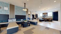 This thoughtfully designed amenity space offers a seamless blend of comfort, functionality, and contemporary style across a single open floor plan. The main lounge area makes an immediate impression with built-in banquette seating upholstered in deep navy, accented by blue and white patterned throw pillows set against a striking textured blue accent wall. Cluster pendant chandeliers cast warm, ambient light over the space, complemented by patterned carpet tiles in varying shades of gray and beige that define distinct zones throughout.

A fully equipped kitchen and dining area anchors one end of the space, featuring a large island with bar seating, herringbone wood-paneled walls, dark cabinetry, stainless appliances, and stylish pendant lighting — ideal for entertaining or casual gatherings. Adjacent to the kitchen, intimate dining booths and café-style seating provide additional options for socializing.

A dedicated game room offers a round poker-style table surrounded by upholstered swivel chairs beneath a bold brass drum pendant light, with curated artwork adorning the walls. Near the large floor-to-ceiling windows, a shuffleboard table adds a recreational focal point to the bright, open lounge.

A secondary lounge area features plush white sofas and side tables, while a café nook near the restrooms includes bistro tables, mixed seating, and a built-in wine rack — completing this versatile and elegantly appointed communal space.
